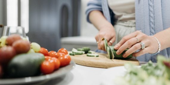 Close-up of a woman slicing cucumbers on a wooden board surrounded by fresh vegetables.