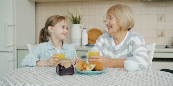 Child and grandmother are clinking glasses with juice and laughing at kitchen table talking having fun together. Family relationship and grandparents concept.